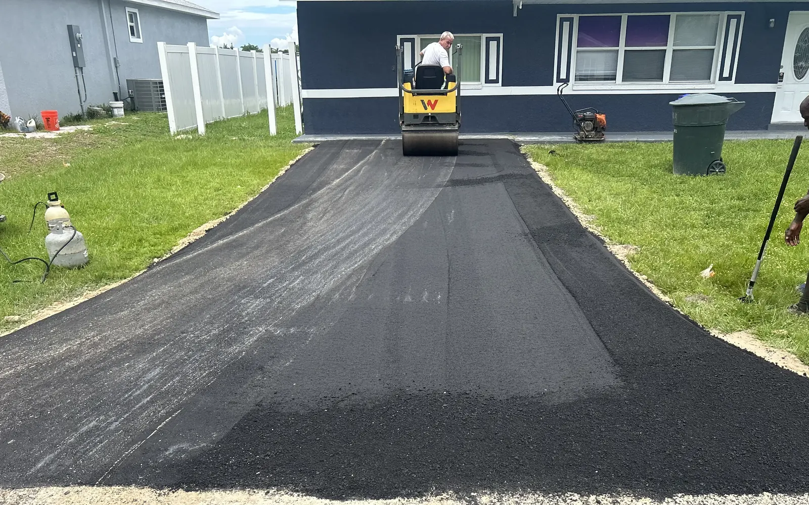 Paving day: the same driveway covered in fresh black asphalt, with a roller compacting it by the house