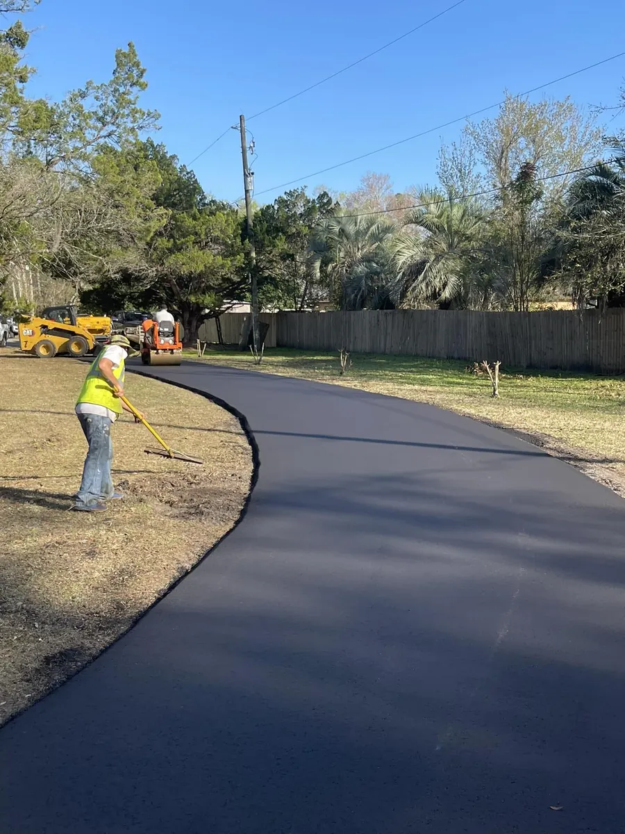 Worker raking the edge of a freshly paved curved driveway, with a roller and cabbage palms behind
