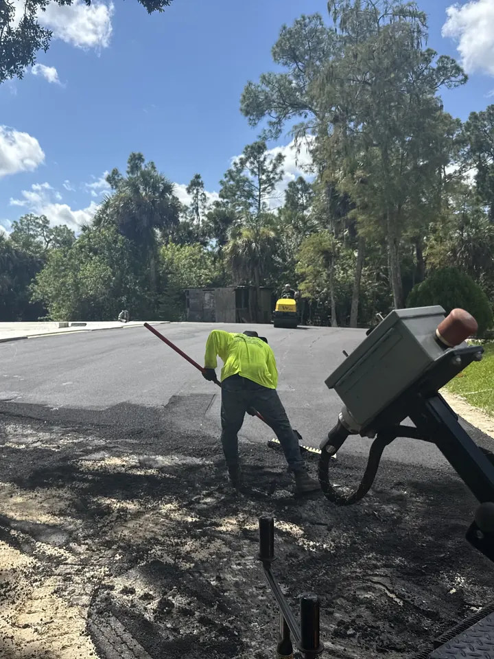 Crew member raking fresh asphalt at the edge of a parking lot, with slash pines and palms behind